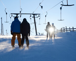 Persons riding a t-bar lift upwards a skiing slope in a winter landscape backlit by the sun shining...