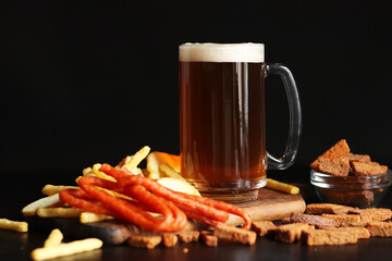 Glass with light beer and snacks on a dark background. Beer, French fries and hunting sausages. Glass on a wooden board and snacks. Light beer and bread croutons. Traditional glass beer mug
