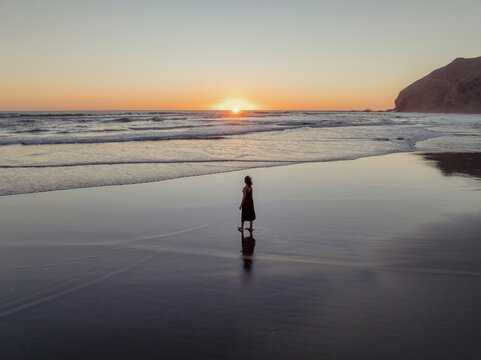 Solitude at sunset. Woman in dark dress walks along tranquil beach at golden hour. Peaceful reflection. Bethells Beach, Auckland, New Zealand