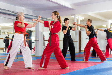 Fototapeta premium Portrait of concentrated elderly woman wearing traditional sportswear sparring with young female opponent during martial arts training in gym..