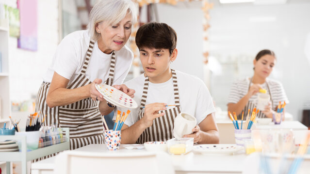 Experienced mature female ceramicist teaching young enthusiastic guy art of ceramic glazing and painting in modern pottery studio..