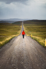 A solitary figure in a red jacket walking on a long, straight gravel road through the remote...