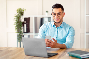 Happy young Arab man in formal wear using smartphone and laptop at workplace. Handsome Eastern entrepreneur having online meeting with partner or colleague, communicating remotely