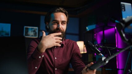 Happy influencer in home studio greeting audience, recording live broadcast using smartphone on selfie stick, close up. Man filming q and a session with mobile phone, answering questions, camera B