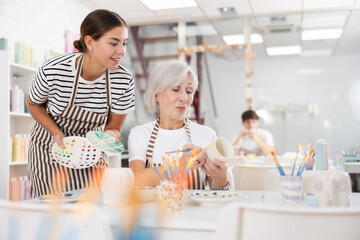 Positive enthusiastic young girl assisting interested senior woman painting ceramic mugs and plates in pottery class. Creative hobby concept..