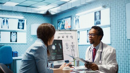 Female patient listening to a diagnostic report from doctor based on MRI scan results and x ray imaging, pointing at computer in medical cabinet. African american expert gives assistance. Camera B.