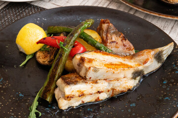 A beautifully plated dish featuring grilled fish, vibrant vegetables, and a wedge of lemon, all served on an elegant black plate.
