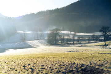 Scenic winter countryside with frosted fields, bare trees, and soft sunlight casting long shadows, surrounded by forested hills.