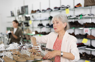 Elderly woman in casual clothes buying flat sandals in shoe store