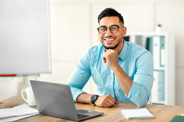 Obraz premium Portrait of happy Arab businessman using laptop computer, making video call, chatting online with colleague at office. Young guy entrepreneur having virtual meeting or business webinar