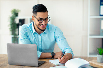 Arab guy entrepreneur writing down information during online business meeting at modern office. Millennial businessman taking notes, watching educational webinar, participating in conference