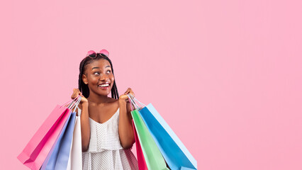 Happy young black lady with shopping bags buying gifts on sale over pink studio background....