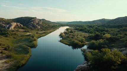 A tranquil river winds through lush greenery and rugged hills under a clear sky, evoking a sense of peace and natural beauty.