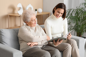 Young nurse with senior woman reading magazine on sofa at home