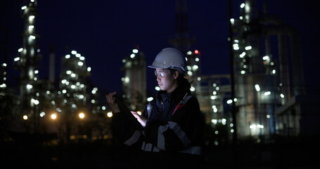 A female chemical worker in safety gear uses a digital tablet at a brightly lit refinery at night