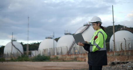 An industry engineer woman using a laptop with a white safety helmet standing in front of the oil refinery. Factory oil storage tank and pipeline. Worker in a refinery