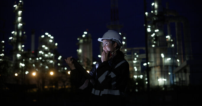A female chemical  worker in safety gear uses a digital  tablet at a brightly lit refinery at night
