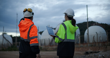 Male Chief engineers with walkie-talkies and female process engineers hold a laptop while working in the chemical storage area field at a petrochemical factory 