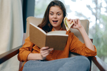 Surprised young woman reading a book while enjoying a sandwich, sitting comfortably in a cozy setting, showcasing joy and relaxation