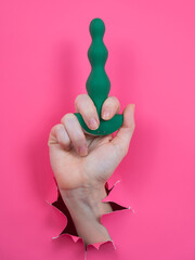 Female hand sticking out with green anal beads from pink paper background. Vertical photo. 