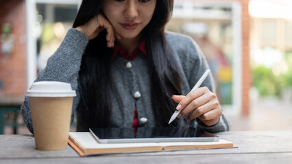 An Asian woman holding a stylus pen, using a digital tablet while sitting at an outdoor table.