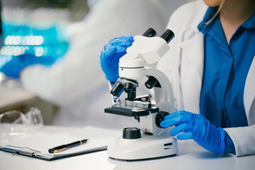 Close-up of scientist's hands in blue gloves adjusting microscope, conducting research in modern laboratory with blurred background and safety goggles on table