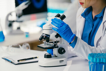 Female scientist wearing gloves using microscope doing scientific research in a modern laboratory with test tubes and computer in background
