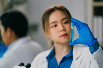 Female scientist wearing blue gloves and a lab coat, holding a test tube filled with blue liquid,...
