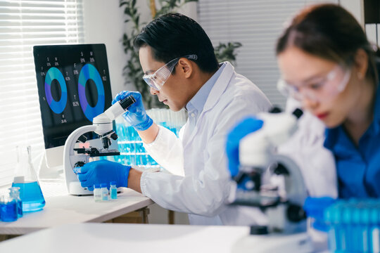 Two medical researchers wearing lab coats and safety glasses are using microscopes and analyzing data on computer screen while developing new treatments in modern laboratory