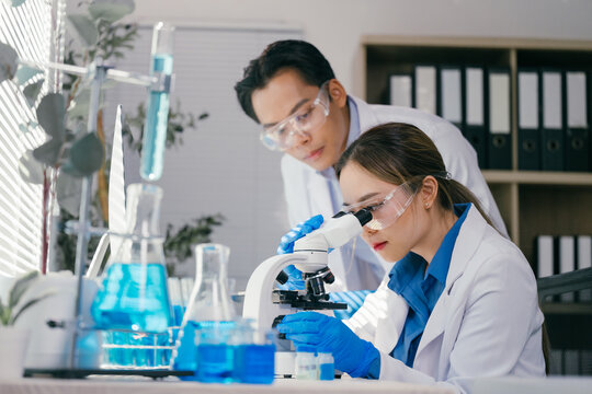 Two scientists in lab coats and safety glasses conducting research with a microscope and test tubes filled with blue liquid for scientific analysis and discovery - Powered by Adobe