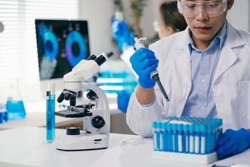 Scientist wearing lab coat and gloves using micropipette dropping blue liquid into test tube doing...
