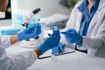 Two scientists wearing lab coats and gloves are examining and comparing samples of blue liquid in...