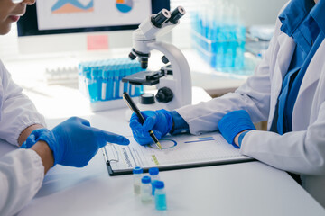Two medical researchers in lab coats and gloves examining data on a clipboard in a laboratory, with a microscope and test tubes highlighting scientific collaboration