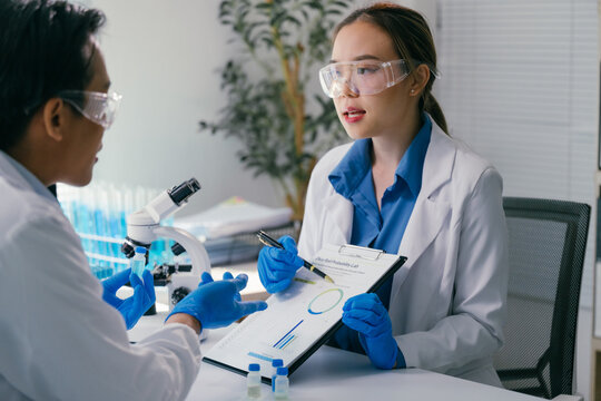 Two scientists wearing lab coats and gloves are examining a document with graphs and charts, discussing research findings in a modern laboratory equipped with a microscope and test tubes