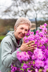 Fototapeta premium A 55-year-old Russian woman enjoying a blooming rhododendron (Ericaceae family) bush in spring. She smiles warmly, touching and smelling the vibrant pink flowers outdoors