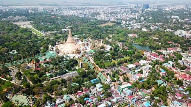 Drone capturing Shwedagon Pagoda, a magnificent golden stupa, surrounded by lush greenery and urban development in Rangun, Myanmar, showcasing the city's blend of religious and modern life