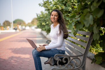 Obraz premium Indian girl, student using laptop at park for education, learning or studying at university on bench in garden. Notebook, serious and woman at outdoor
