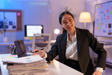 Young professional woman sitting at her desk late at night working on a laptop and holding a pen while reviewing paperwork with a large stack of files next to her