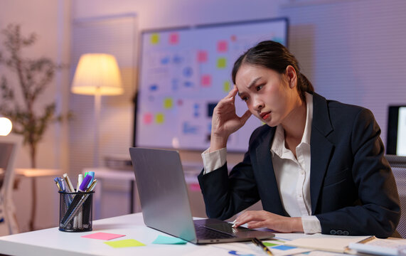 Young manager experiencing a headache while working late at night in the office, touching her head and displaying signs of stress and fatigue while focused on her laptop