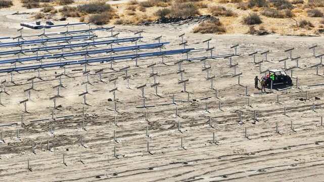 Workers driving an ATV through a desert solar panel construction site, surrounded by rows of metal framing structures under a bright sunny sky. TRUCKING RIGHT TELEPHOTO