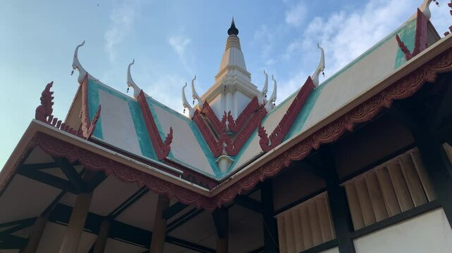 A decorative Cambodian white temple roof with finial and chofah stands against a blue sky with white clouds, low angle