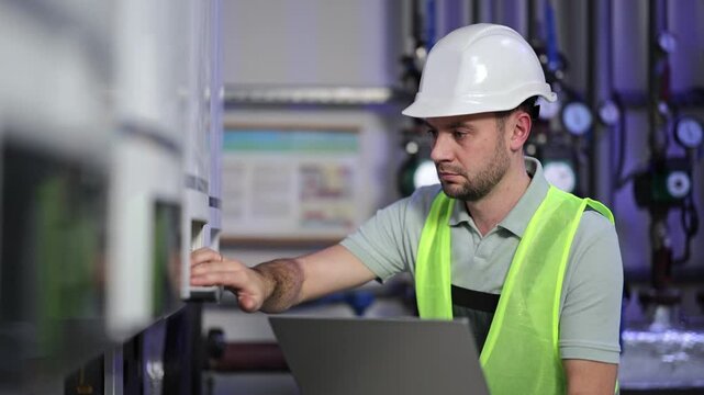 Serious, focused power generation plant worker types data into laptop. Industrial environment with equipment and gauges in background. The competent male engineer monitors operations.