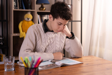 Thoughtful teenage boy reading book at desk in study area - educational concept.