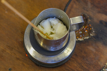 Close-up of an iron bowl on a stove where beeswax is melted to make candles. The woman stirs the wax periodically. The process of making a handmade scented candle in a home workshop.