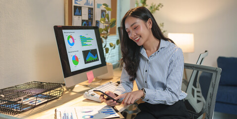 Young businesswoman smiling and analyzing charts and graphs using smartphone and computer screen, working on financial data in modern office