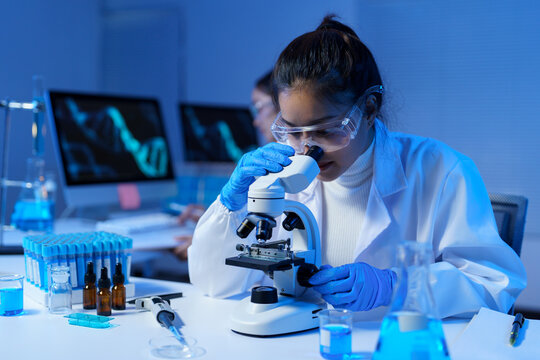 Young female scientist wearing a lab coat and gloves analyzing samples with a microscope in a modern laboratory, with blue light and computer screens displaying dna structures