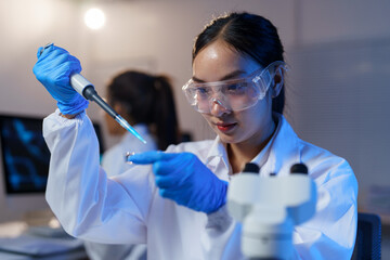 Asian female scientist wearing protective glasses and gloves using micropipette dropping liquid...
