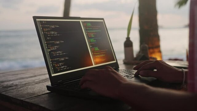 Software developer types on laptop at beachside cafe, ocean sunset backdrop. Male programmer writes code, working remotely near palm trees, sea waves during golden hour. Freelancer project coast.