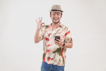 Happy young Asian man holding his smartphone while showing an okay finger gesture.