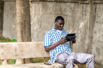 Obraz premium African American man seated in the garden calmly using phone for leisure, Technology, game, gamer, gaming, connection